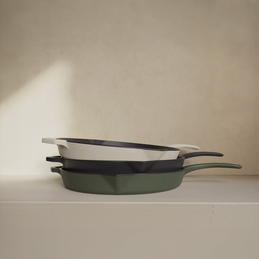 Three enamelled cast iron pans in white, black, and green are stacked on a light surface in soft natural light.